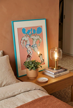 Load image into Gallery viewer, Another Day, Another Modelling Gig!! by IsaLu Ishii – colourful art print featuring two cowboys with blue hats, shown framed, leaning against a wall on a bedside table.
